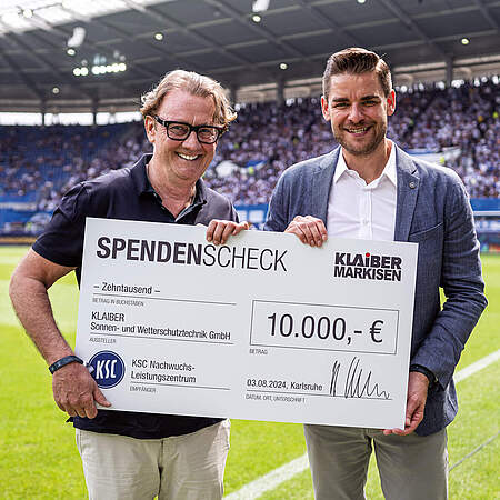 Ralf Klaiber und Michael Becker halten einen großen Scheck über 10.000 für die KSC aKAdemie in der Hand.